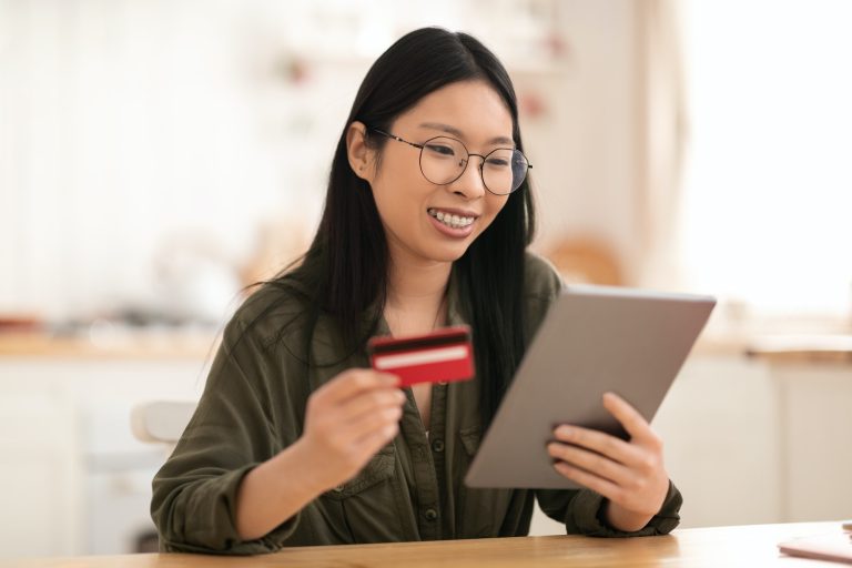 Smiling asian woman using tablet and bank card at kitchen